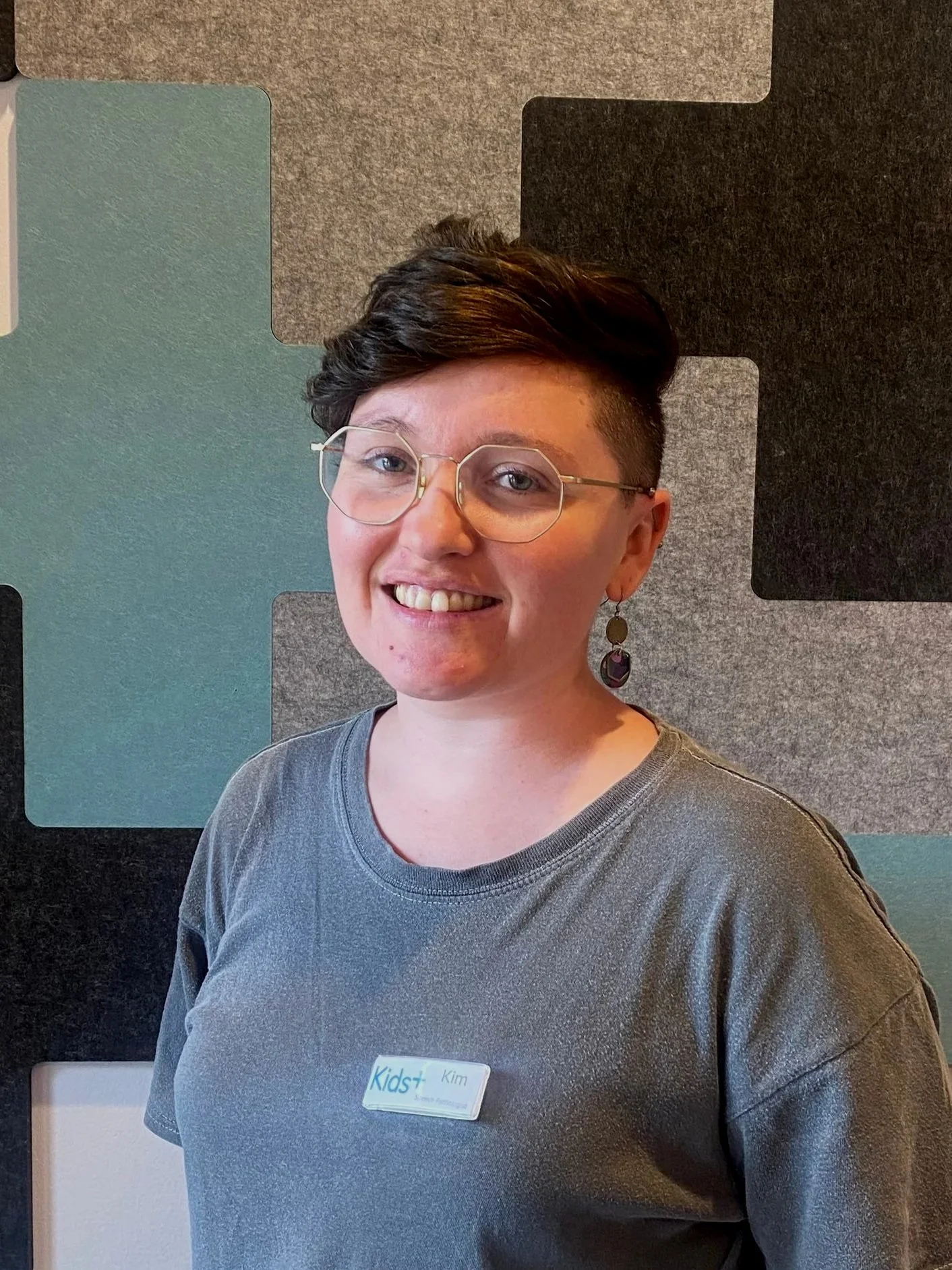A smiling woman wearing glasses and a gray t-shirt, standing in front of a colorful geometric-patterned wall.