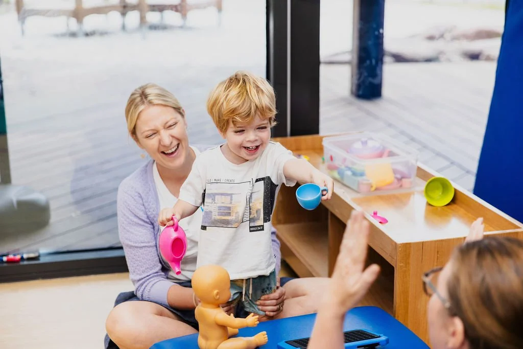 smiling blond young boy offers a plastic blue teacup to a therapist as a second therapy supports his legs from behind