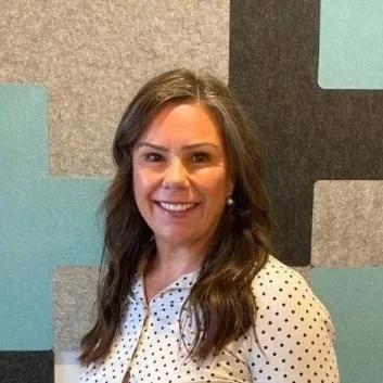 Headshot of a woman with long brown hair wearing a white blouse, smiling, standing in front of a geometric patterned background.