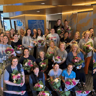 large group of smiling women holding bouquets of flowers in front of an illuminated kids+ wall sign