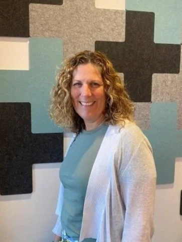 A woman with curly blonde hair smiling, standing in front of a wall with geometric colored panels.
