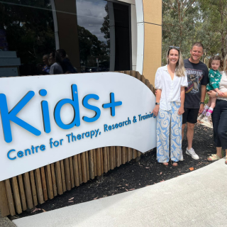 Group of three adults and one child standing outside near a sign that reads 'Kids+ Centre for Therapy, Research & Training' in front of a building.
