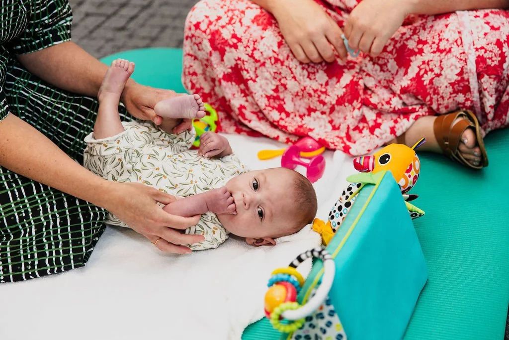 A baby lying on a changing table with a caregiver holding its leg, and another person sitting nearby. The baby has a thoughtful expression and is sucking on a thumb. Colorful toys surround the baby, including a giraffe and patterned plush toys.