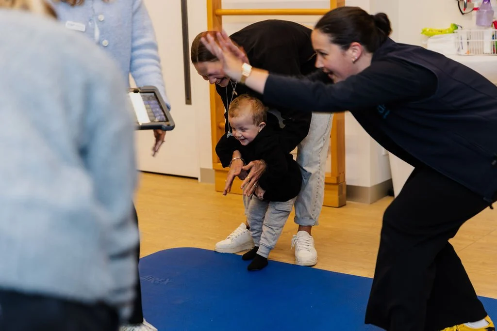 A young child in black clothing laughing and smiling as an adult assists him during a physical therapy session on a blue mat, with another adult and a person holding a tablet nearby.