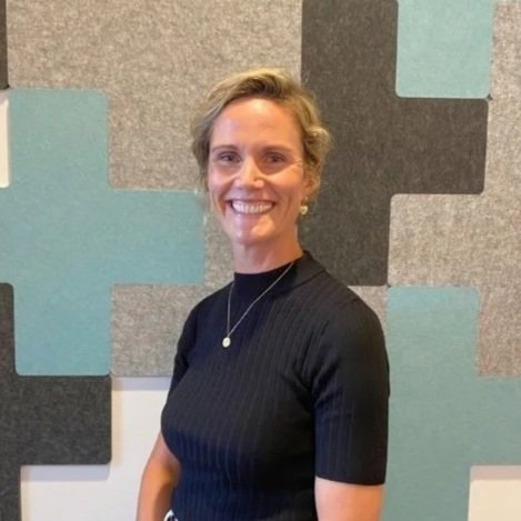 A woman with short blonde hair, wearing a black ribbed top and a necklace, smiling in front of a gray and teal patterned background.