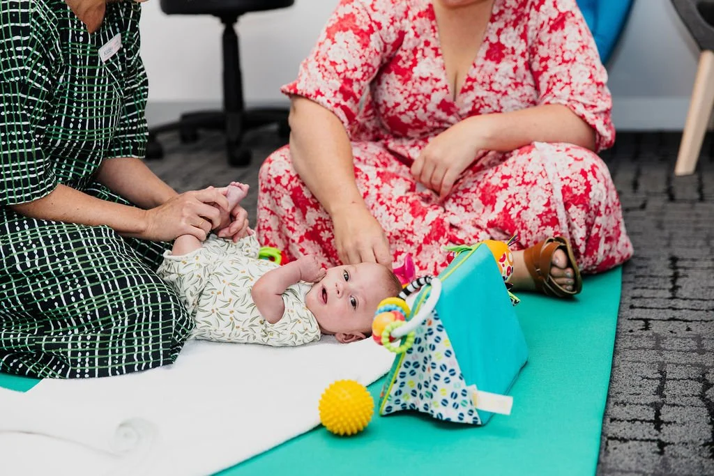 Two women sitting on a turquoise mat with a baby lying on its back. One woman is holding the baby's hand, and the other woman is reaching towards the baby's head. The baby is wearing a patterned shirt, and there are colorful toys around them.