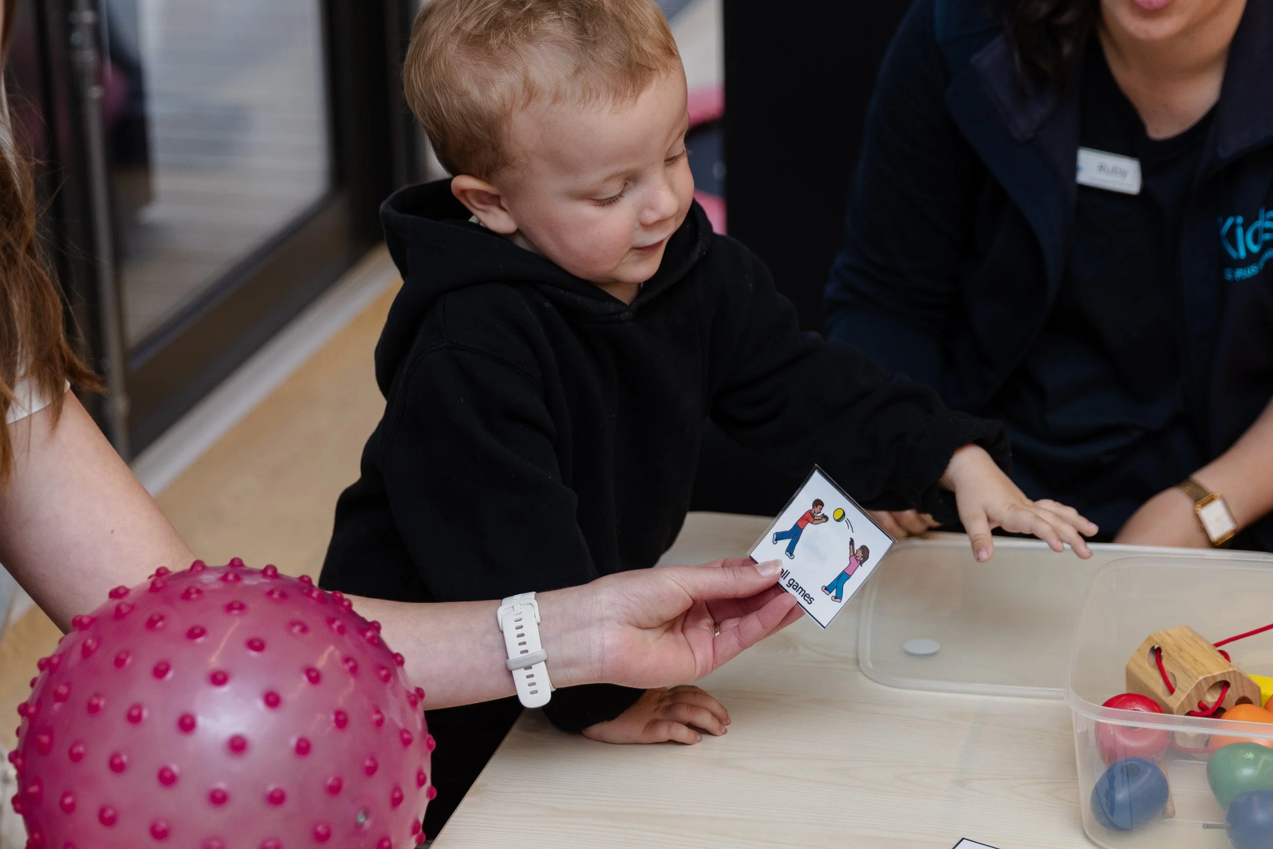 A young boy with red hair and a black hoodie playing a game with a card showing an illustration of two children playing tennis, held by an adult woman with a white wristwatch. Other children, adults, and toys are visible in the background.