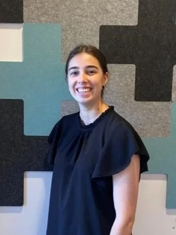 A woman with brown hair, wearing gold hoop earrings and a black ruffled blouse, standing in front of a geometric patterned wall.