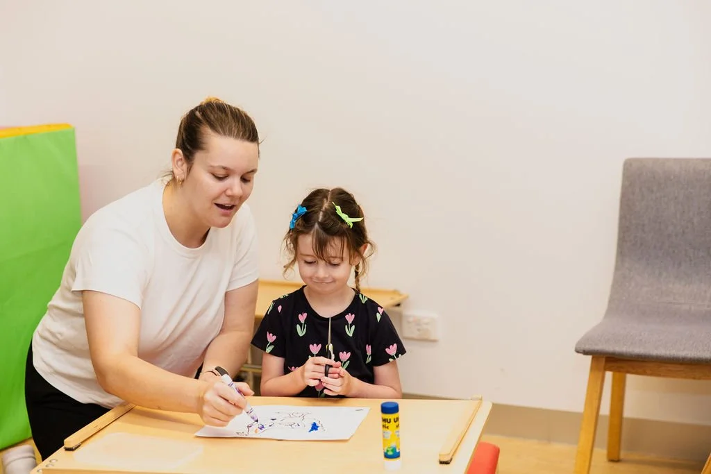 A woman and a young girl drawing together at a small table in a room with a plain wall, a gray chair, and colorful foam blocks in the background.