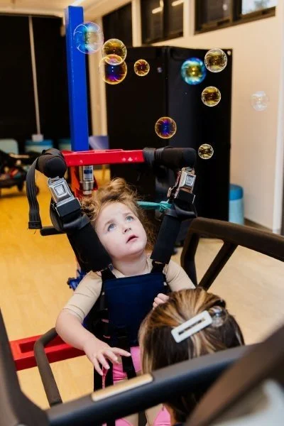 young girl in body support harness gazes up at bubbles during a physical therapy session.