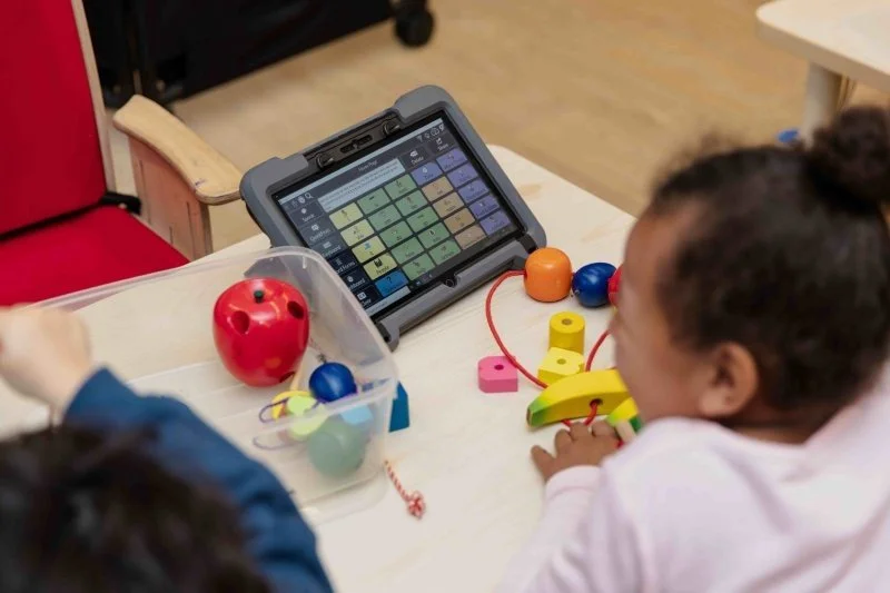 young girl using an aac device surrounded by toddler toys