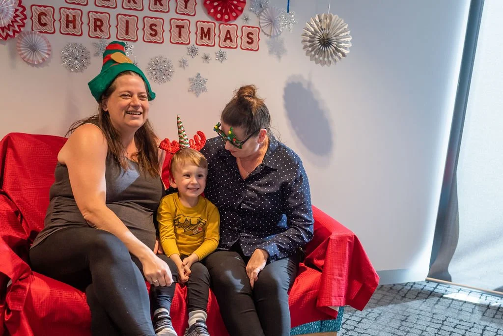 Three people sitting on a red couch decorated with Christmas items; a woman wearing a green elf hat, a young boy with reindeer antlers, and a woman with holiday glasses and reindeer headband, celebrating Christmas indoors.