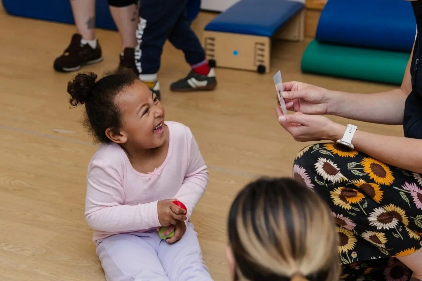 A young girl sitting on a wooden floor, laughing and holding a small ball, during a playful activity with an adult woman showing her a card, in a room with gym mats and other children in the background.