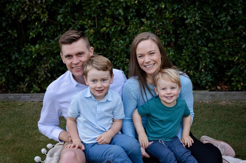 father and mother and two young boys sit in front of a green hedge and smile broadly