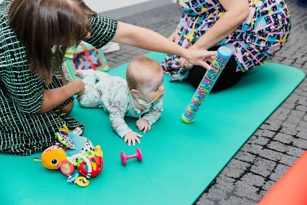 baby playing with toys on colourful mat supported by two adults
