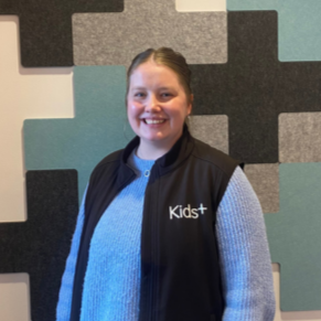 A young woman with light brown hair in a ponytail, smiling, wearing a blue sweater and a black vest with the logo 'Kids+' on it, standing in front of a background with colorful acoustic panels.