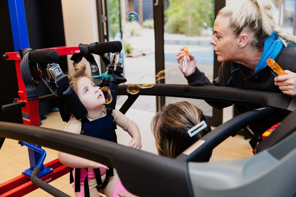women blows bubbles for young girl who sits in a harness on a treadmill. a physiotherapist is holding the girls legs