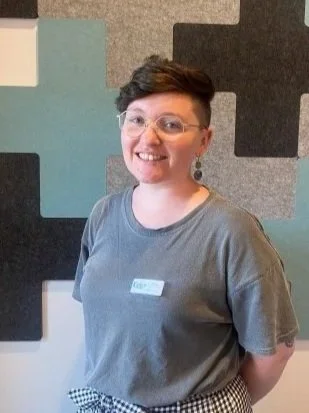 A woman with short dark hair, glasses, and earrings smiling in front of a geometric patterned wall, wearing a gray T-shirt with a name tag that reads "Kids* Kim".