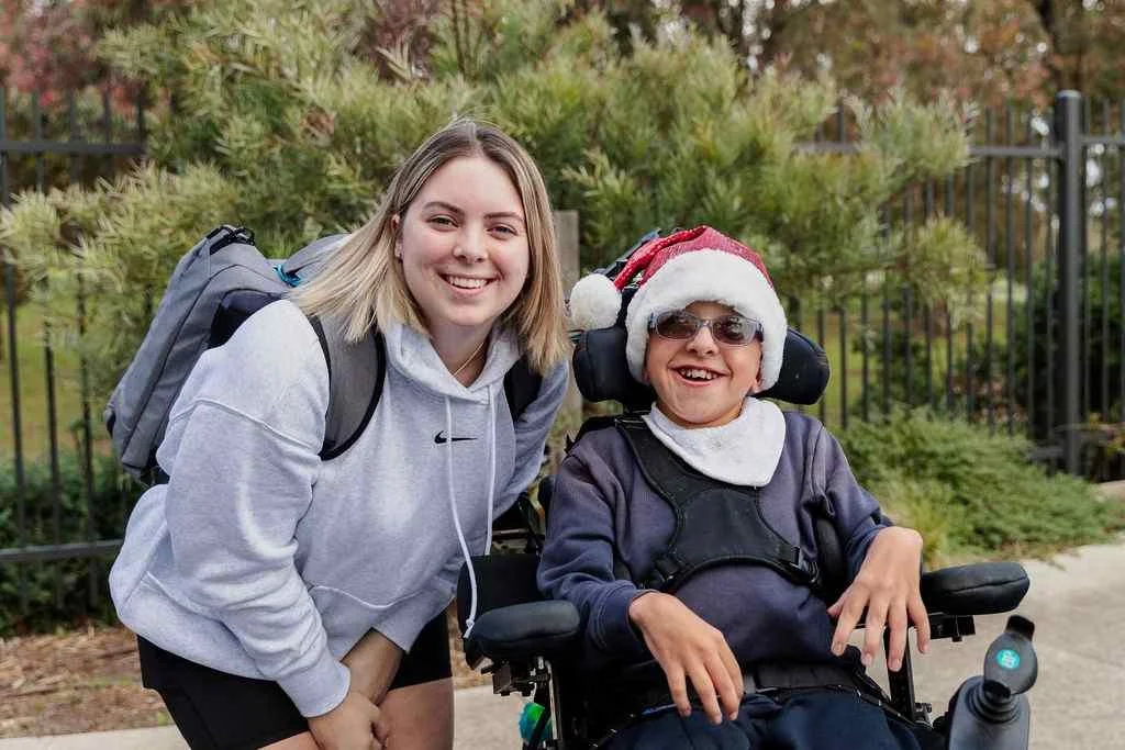 young boy in wheelchair wearing a santa hat smiles beside a woman