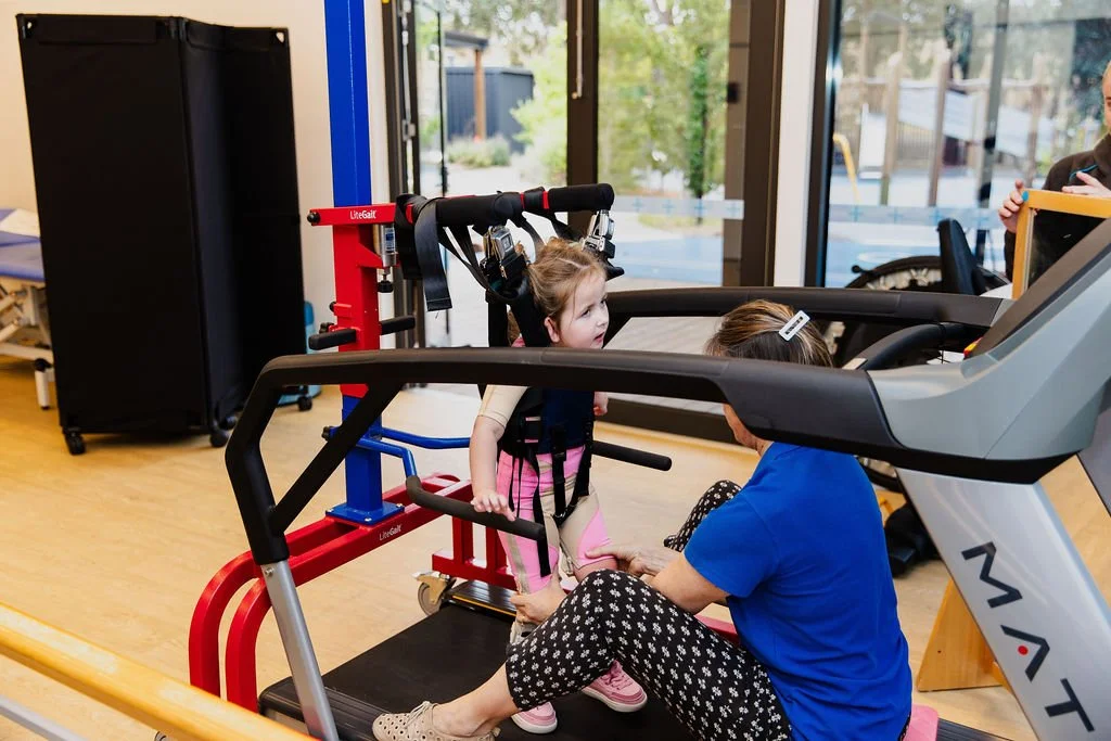 A young girl in a harness and pink leggings standing on a treadmill with a woman in a blue shirt assisting her in a therapy or rehabilitation setting inside a facility with large glass doors and outdoor view.