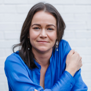 A woman with shoulder-length dark brown hair and light skin, wearing a blue blouse, sitting with her arms crossed and resting on a wooden chair, smiling gently in front of a white brick wall.
