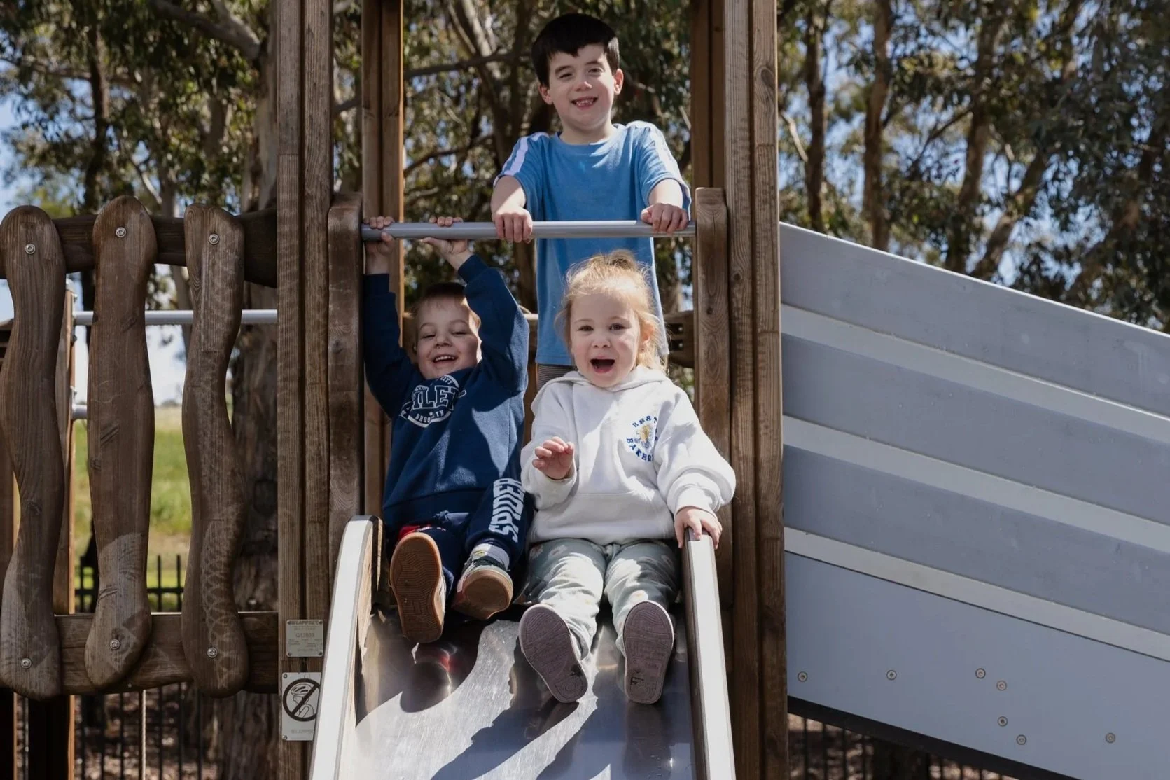 Three children playing on a playground slide, with trees in the background on a sunny day.