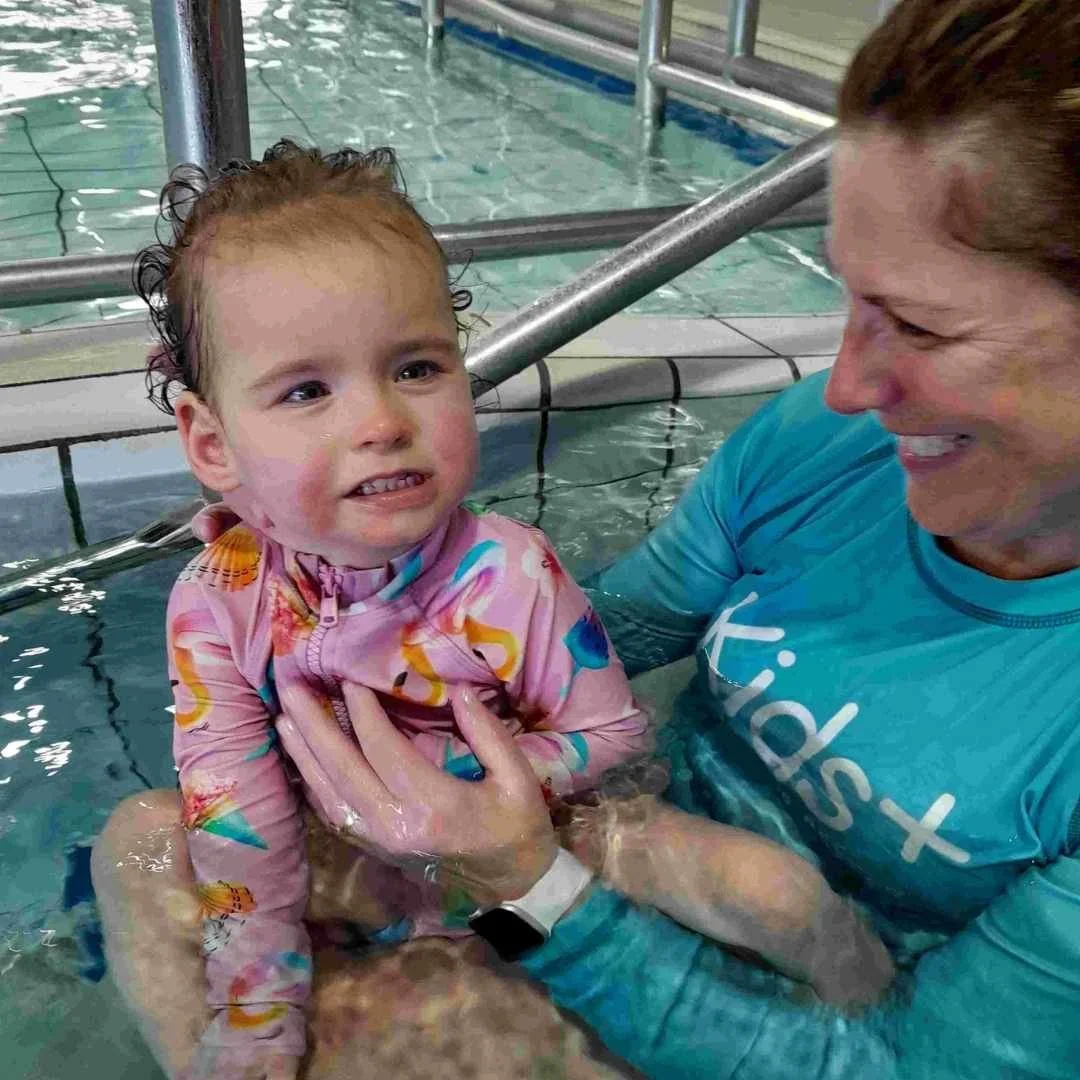 young girl in swimming pool smiles at camera. she is bring held upright by a therapist