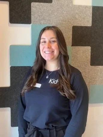 A smiling woman with long brown hair, wearing a black Kids shirt, standing in front of a colorful wall with square patterns.