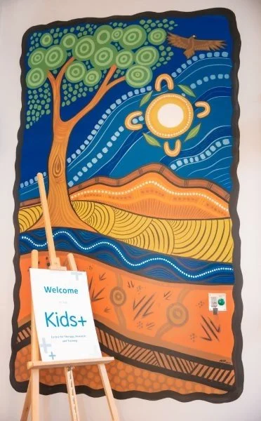 aboriginal painting on a wall with wooden a-frame holding a Kids+ blue logo on white card