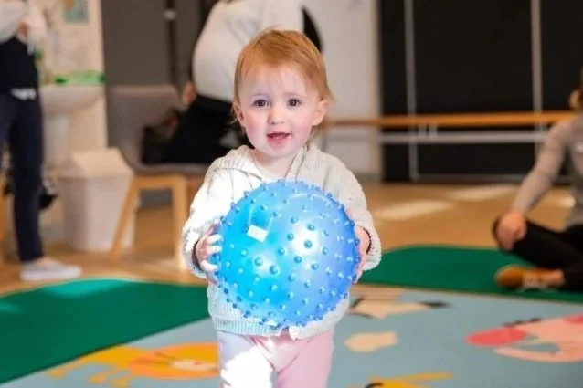 young toddler holds soft, spikey, blue ball and smiles