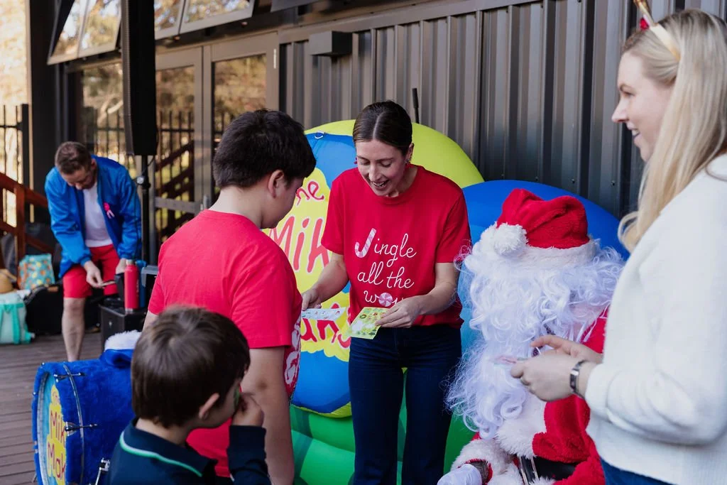 A group of people, including children and adults, exchanging gifts at a holiday event outdoors. Santa Claus is present, wearing his red suit and Santa hat, sitting among large colorful balloons with holiday lettering.