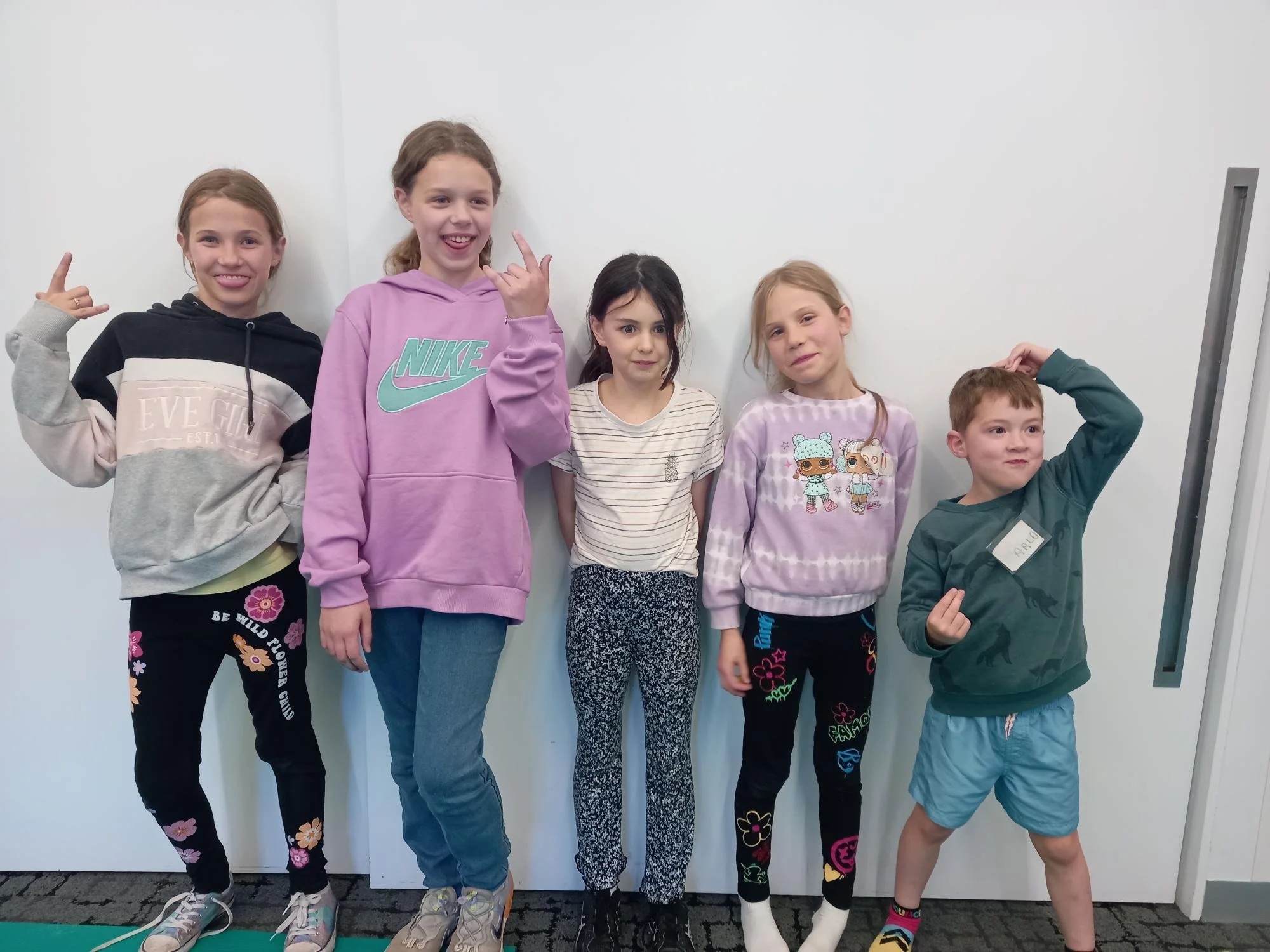 Group of five kids standing against a white wall, posing for a photo, with some wearing colorful casual clothes and sneakers.