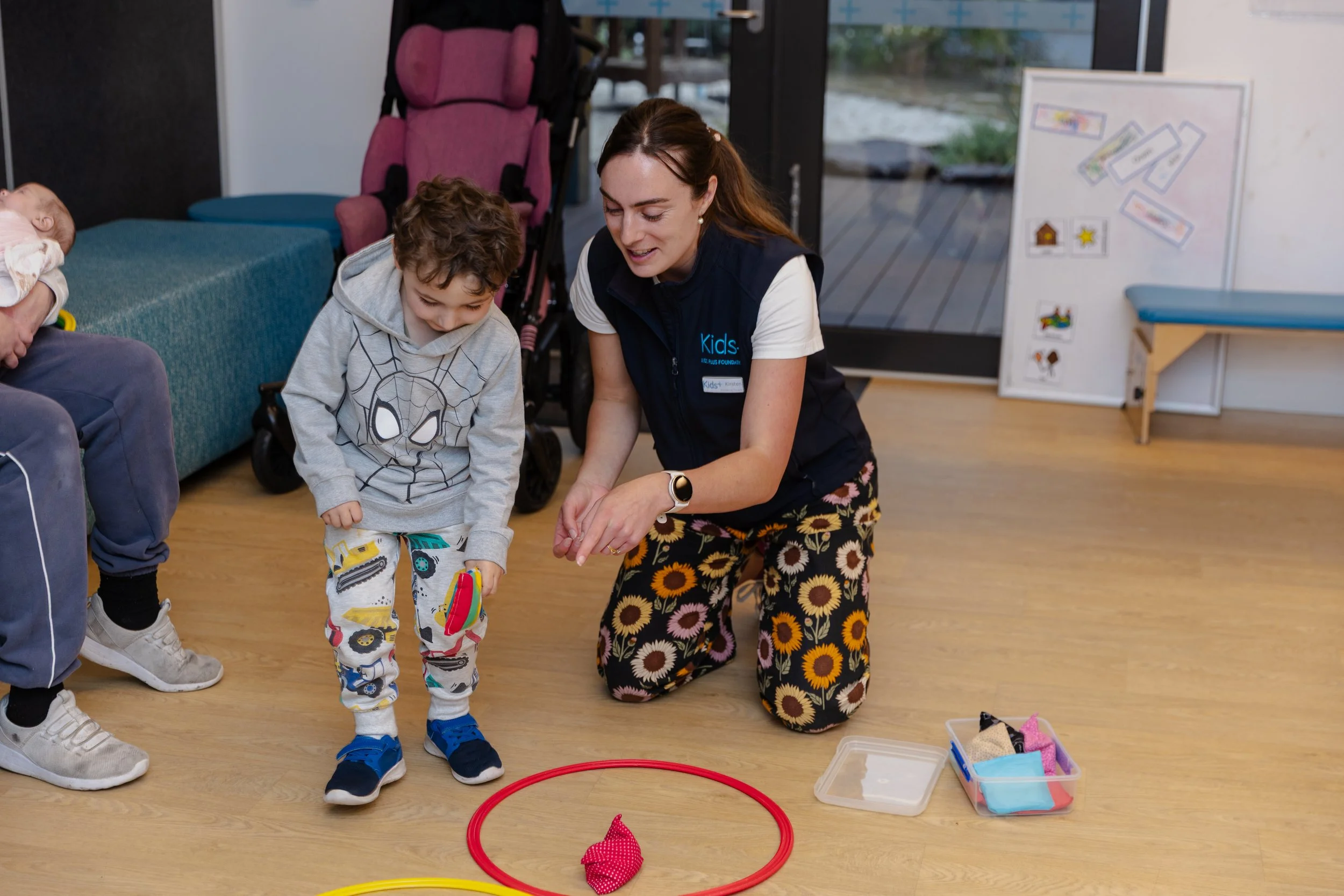A woman kneeling on the floor with a child, participating in a sensory play activity with a red hoop and a small fabric bag, in a room with other children and adults.