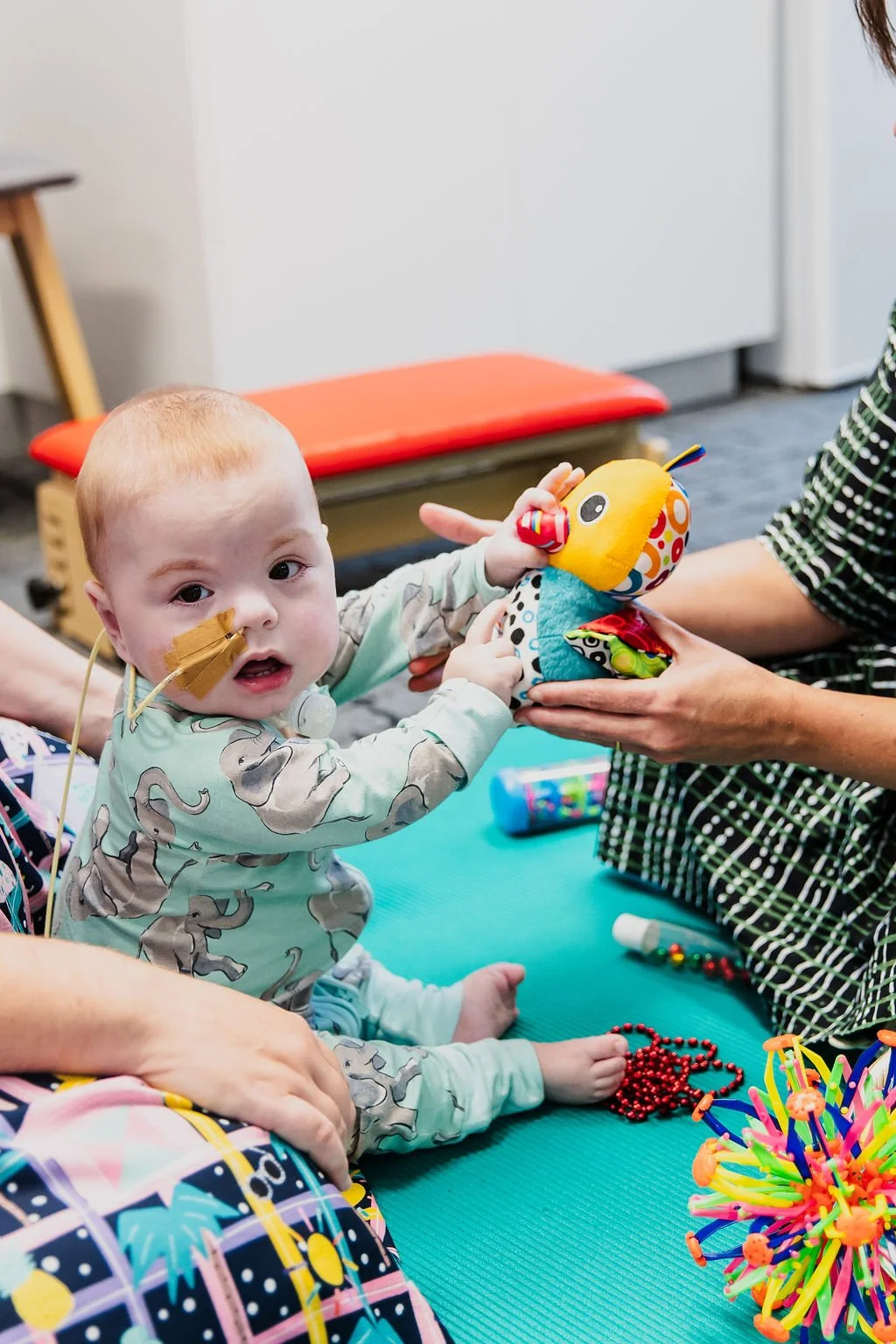 A young child with a nasal tube and hospital gown sitting on a blue mat, holding a colorful plush toy, while an adult helps hold the toy during a therapy or play session.
