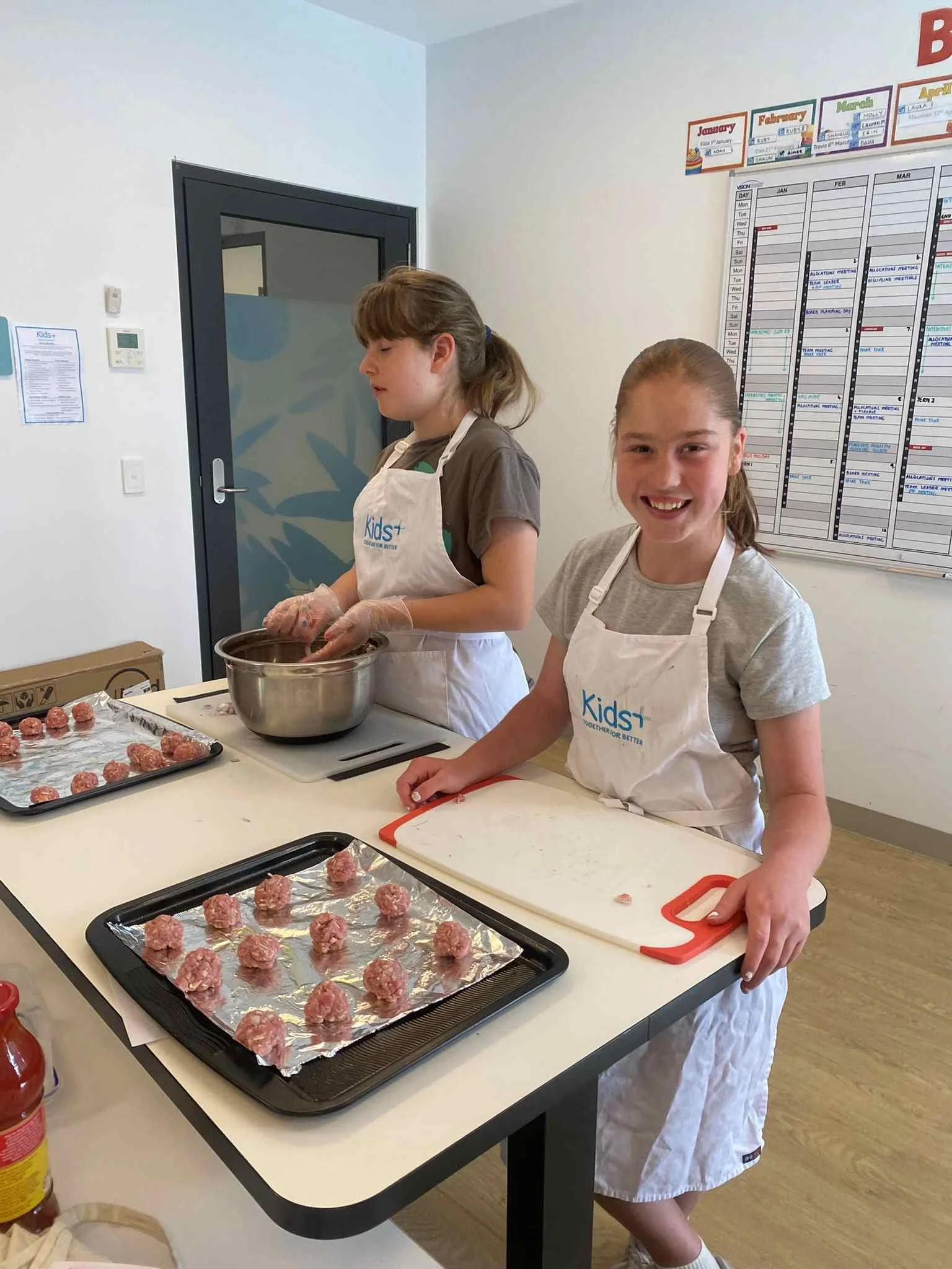 two young girls baking meatballs wearing kids+ white aprons smiling