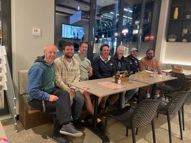 Seven men sitting at a dining table in a restaurant, smiling for the photo.