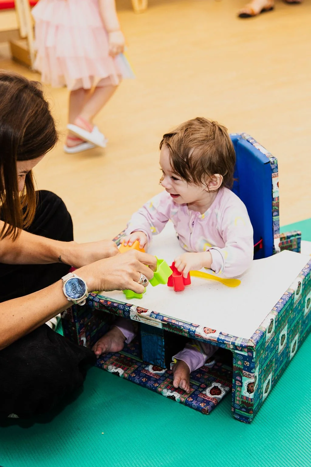A young girl sitting in a high chair smiling as an adult hands her colorful toy shapes, with others in the background.