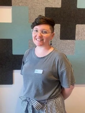 A smiling woman wearing glasses and a gray t-shirt, standing in front of a colorful geometric-patterned wall.