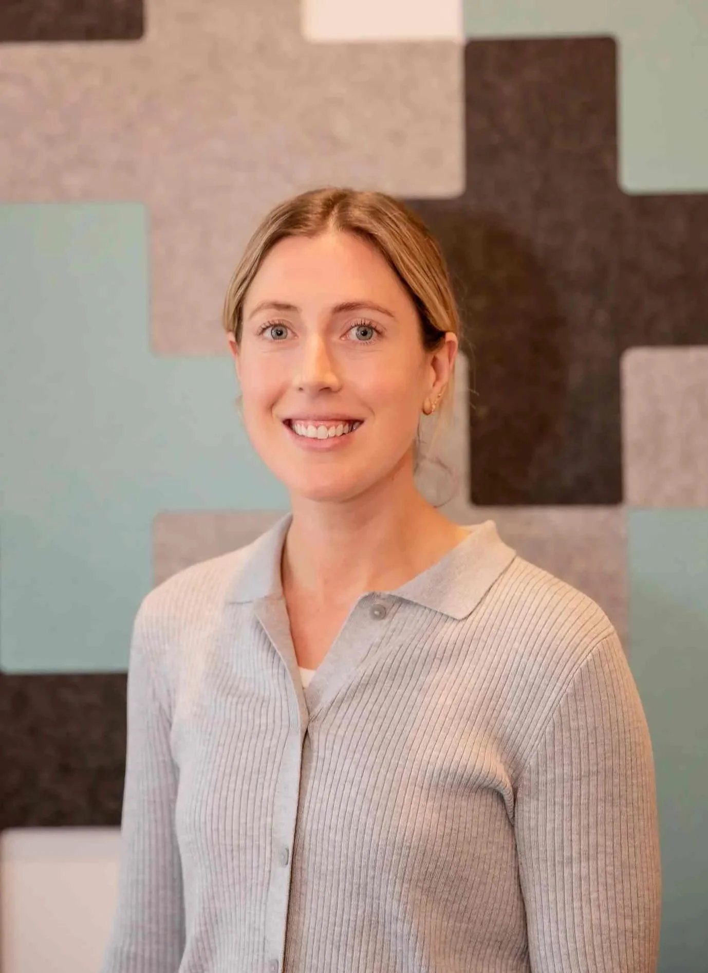 A woman with blonde hair, blue eyes, and a friendly smile, wearing a light gray, ribbed, long-sleeve shirt with a collar, standing in front of a colorful, textured wall with abstract geometric shapes.