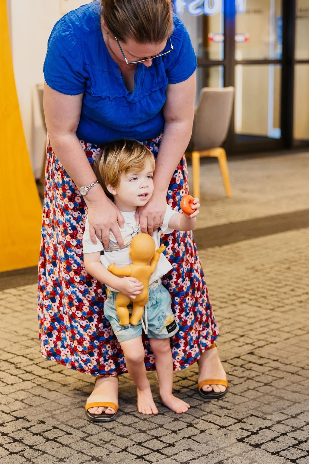 A woman in a blue top and floral skirt helping a young boy, who is barefoot and holding a toy doll and a small rubber duck, stand on a carpeted floor inside a building.