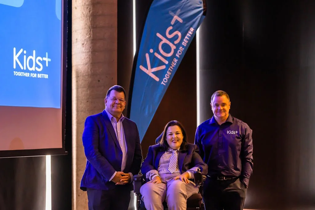 Three people posing together at a Kids+ event, with a large screen and a blue Kids+ banner in the background. The woman in the middle is in a wheelchair and all three are smiling.