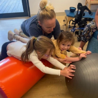 A woman and two young girls engaging in physical therapy with a large orange foam roll and a gray exercise ball on a wooden floor inside a room with a door and equipment in the background.