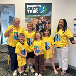 4 children and 2 adults in matching yellow printed tshirts pose in front of a surf coast trek event poster