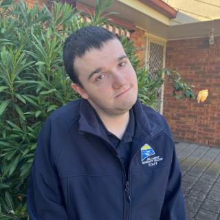 young man smiles at camera in front of a green bush outside a red brick house