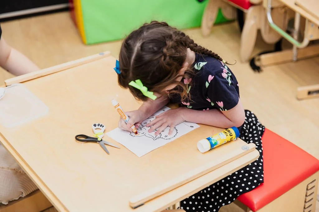 A young girl with braided hair and colorful bows coloring on a sheet of paper at a classroom table. Art supplies like scissors, glue, and markers are on the table.