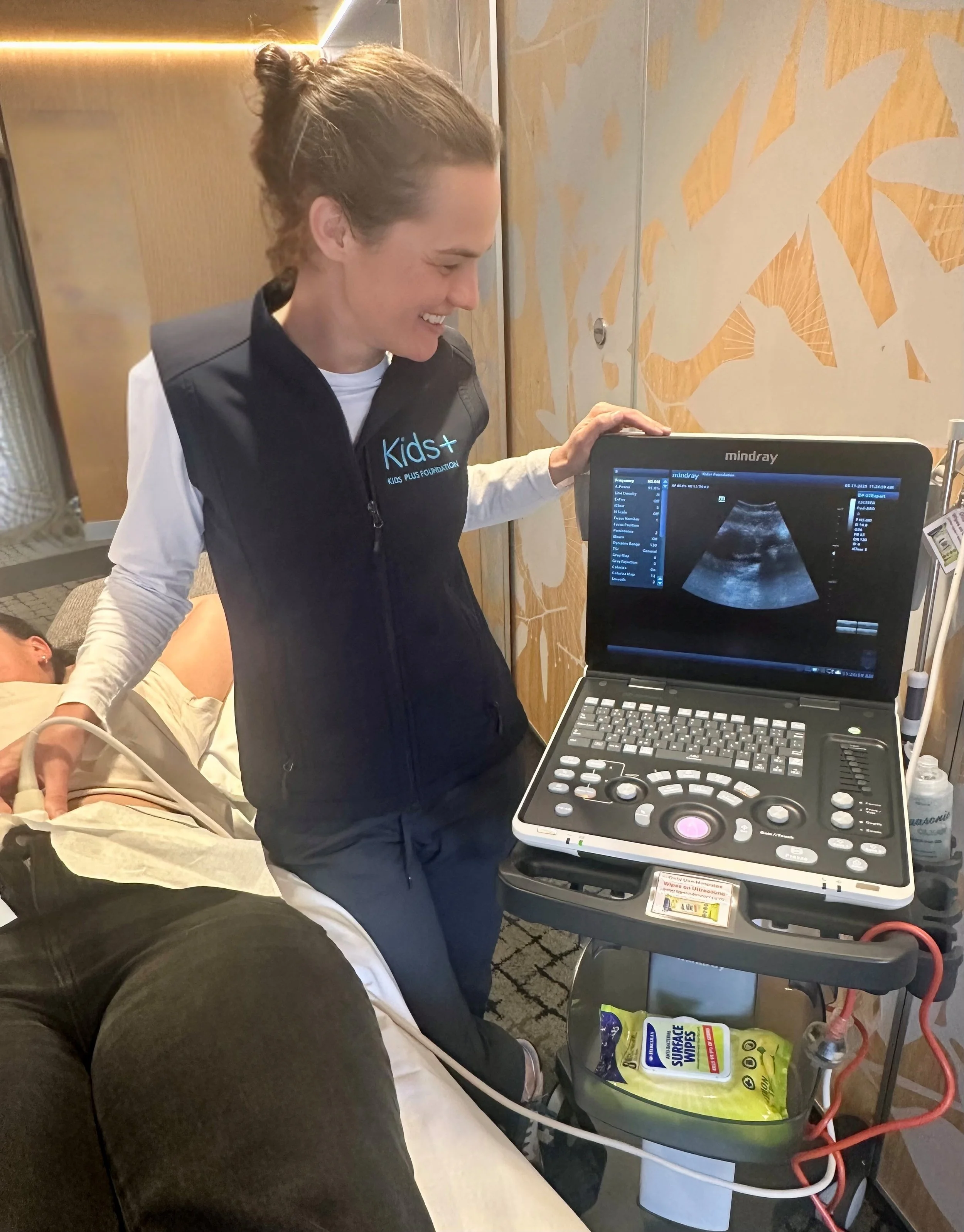 A woman smiling while using an ultrasound machine on a person lying down on a medical bed.
