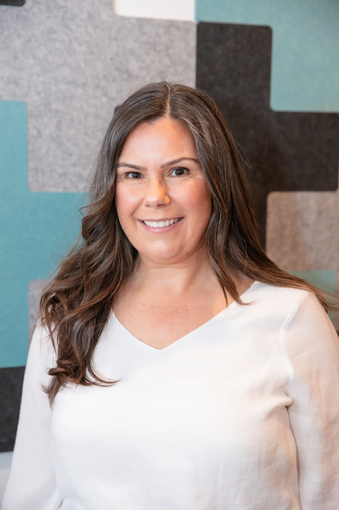 Headshot of a woman with long brown hair wearing a white blouse, smiling, standing in front of a geometric patterned background.