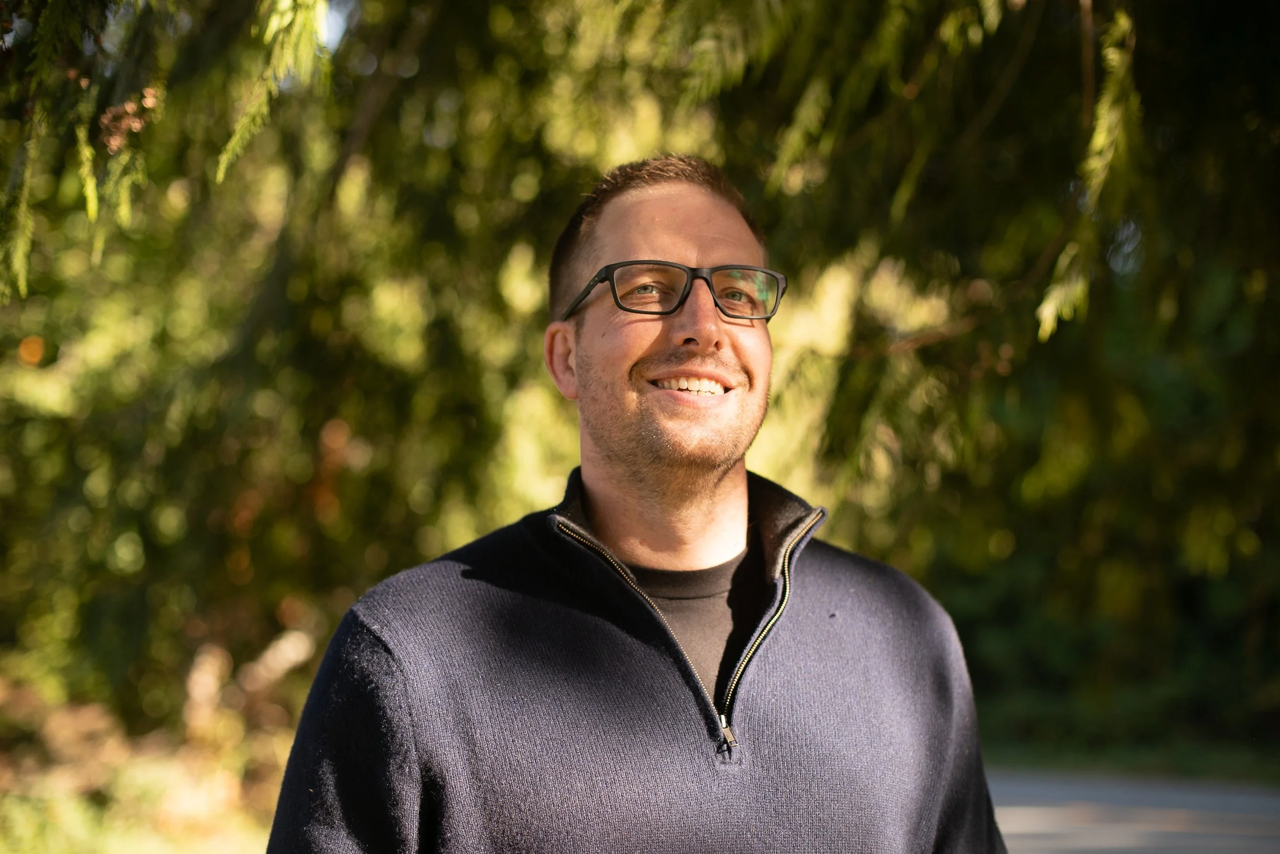 A smiling young man with light brown hair standing outdoors by the water on a cloudy day.