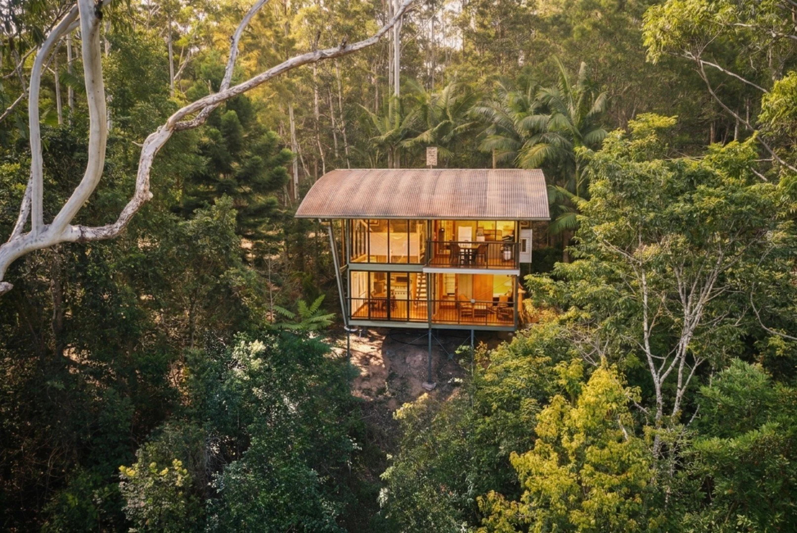 A modern glass house built on stilts on a hill, surrounded by dense green trees, with warm interior lighting.