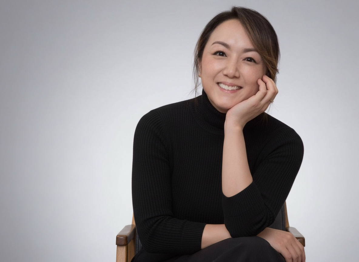 A woman with brown hair in a black turtleneck sweater smiling and resting her head on her hand, sitting on a wooden chair against a plain white background.